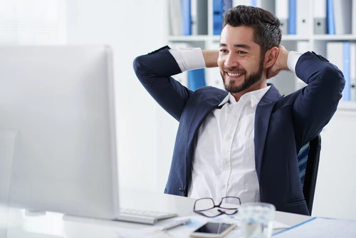 Homme d'affaires souriant en costume bleu marine et chemise blanche, mains derrière la tête en position de détente dans un bureau moderne